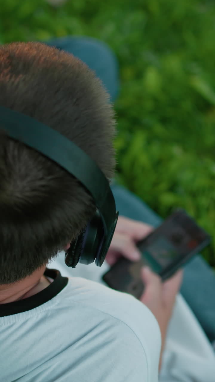 vista trasera del productor de música con auriculares, asintiendo con la cabeza a la música mientras opera su teléfono, está sentado en un campo cubierto de hierba rodeado de vegetación vibrante y luz natural