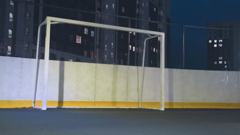 soccer ball kicked into goal post bounces back and rolls out on an urban sports field at night, illuminated by artificial lighting, with background featuring residential buildings and safety netting