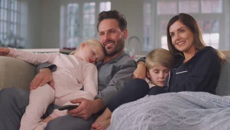 family in pyjamas sitting on sofa watching tv together as children fall asleep