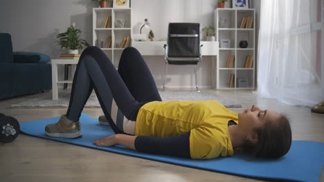 Ejercicio-Físico-En-Casa-Una-Mujer-Joven-Está-Entrenando-Sola-En-La-Sala-De-Estar-Por-La-Mañana-Tumbada-En-El-Suelo-Y-Levantando-Las-Caderas-Tensando-Los-Músculos-Abdominales-Y-De-La-Espalda