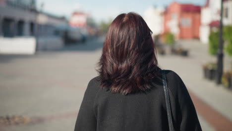 close-up back view of a woman with brown hair walking in a black coat, captured on a sunny urban street, showcasing everyday elegance