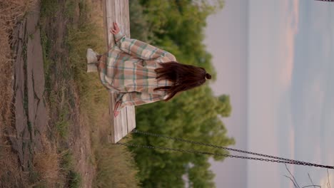 Vertical-video:-A-brunette-in-a-checkered-shirt-swings-on-a-wooden-swing-outside-the-city-on-the-weekend.-Rest-in-the-country-by-the-sea