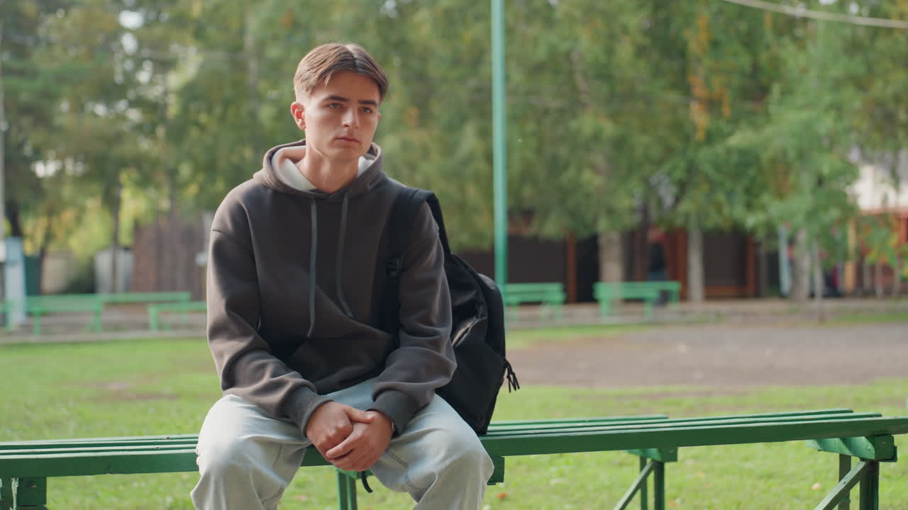 Bílý dospívající sportovec sedí na zelené lavičce na sportovním hřišti, má na sobě mikinu a batoh, ruce sepnuté, pohled do dálky, prázdný pavilon za ním, teplé filmové tóny, dokumentární styl zachycující nervózní očekávání