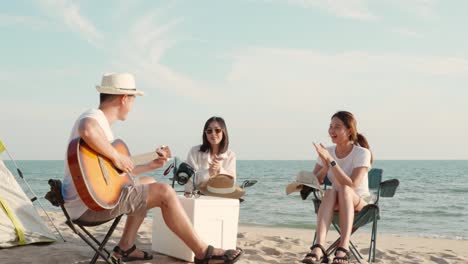 행복한 친구는 캠핑 텐트 근처의 모래 해변에서 휴가를 함께 미소 짓고 캠핑에서 기타를 연주하고 박수를 치며 즐거움을 얻습니다.