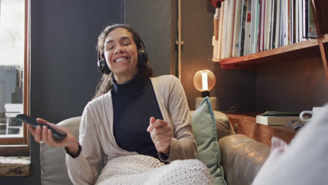 biracial woman lying on sofa under blanket listening music using smartphone at home, slow motion
