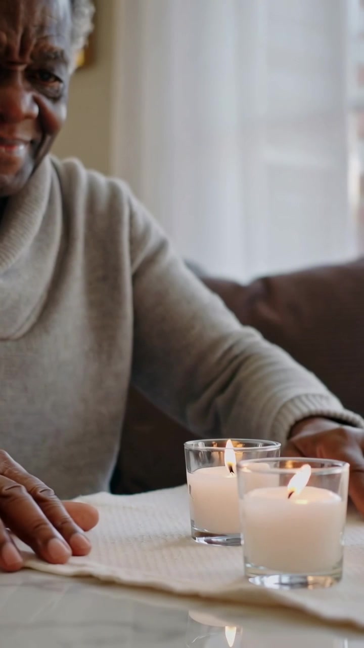 Vertical video: Smiling man in sweater placing two lit votives on home table for calm, copy space