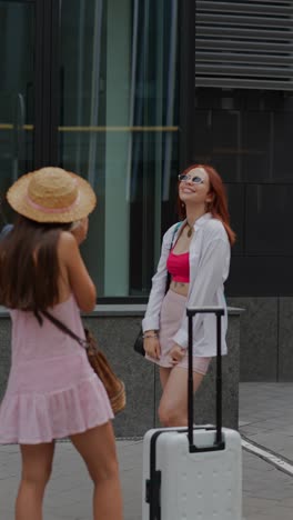 two women taking photos and posing with suitcase outside building