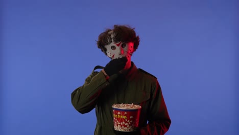 man in white mask eating popcorn on blue background with red neon light image of maniac jason - man in white mask eating popcorn on blue background with red neon light. image of maniac jason voorhees from friday 13. halloween celebration concept