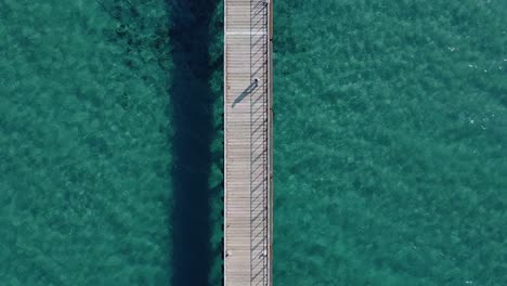 overhead drone view of a person walking along a long wood pier surrounded by sparkling tropical waters