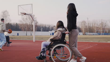 Behinderte-Frau-Im-Rollstuhl-Und-Ihre-Freundin,-Die-Ihren-Freunden-Zusieht,-Wie-Sie-Basketball-Spielen-1