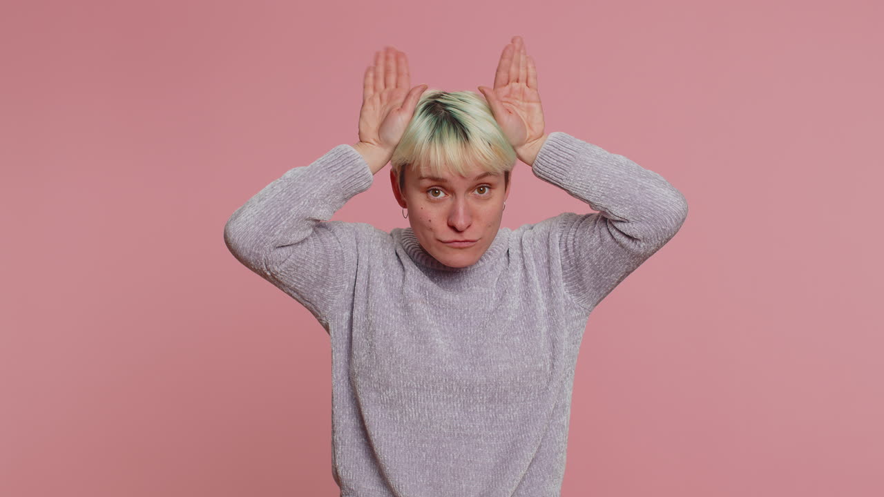 짧은 머리카락을 가진 사랑스러운 재미있는 여성은 친절하게 미소 짓고 머리에 토끼 귀의 제스처를하고, 속이고