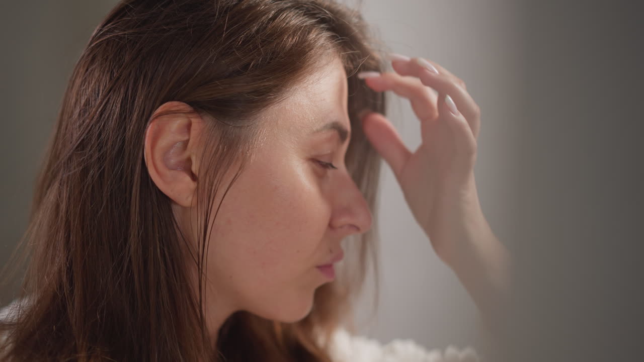 머리카락의 질을 검사하는 거울을 보는 여성. 벌거벗은 매니큐어를 가진 갈색 여성은 화장실에서 귀 근처의 피부를 진지하게 느린 동작으로 봅니다.