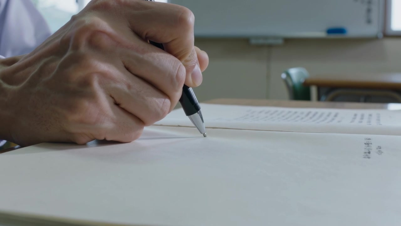 Reading printed list, right hand lowering black pen marking notebook in classroom, pale shirt cuff