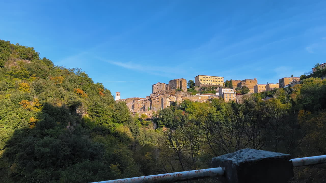 Weitwinkelaufnahme einer italienischen Altstadt auf einem Hügel, umgeben von grünen Wäldern und blauem Himmel – aus der Perspektive eines Autos.