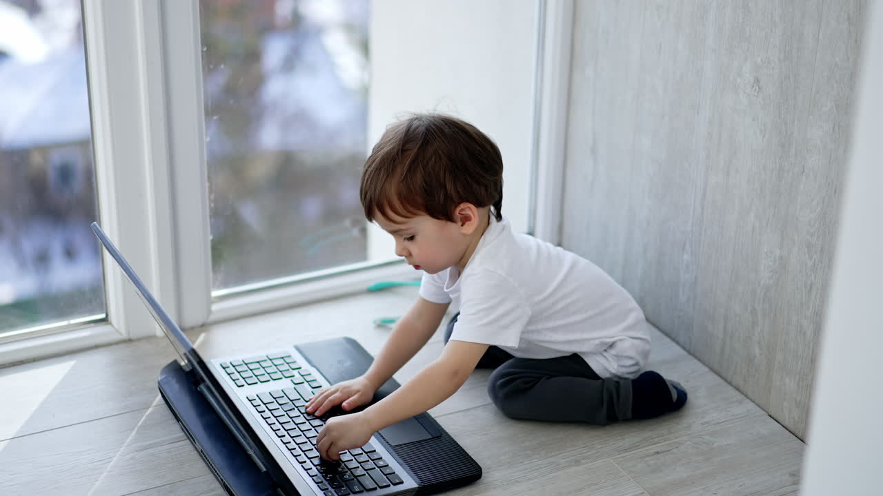 Cute dark-haired boy sits on the floor near laptop. Interested toddler carefully presses the keys on the keyboard. Top view.