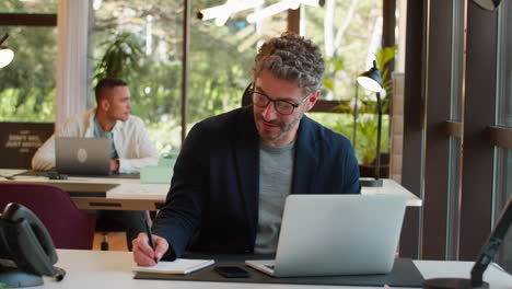 Mature-Businessman-Wearing-Glasses-Working-In-Office-Writing-In-Notebook