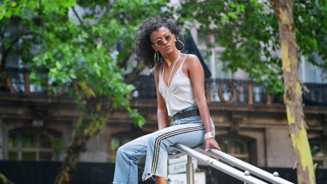 trendy young black woman wearing camisole and side stripe jeans, sitting outdoors on handrail