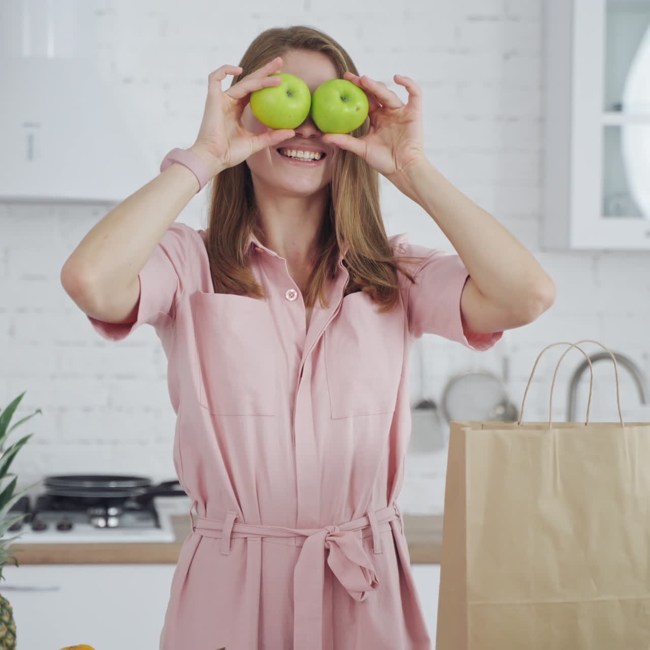 Beautiful woman in light dress holding green apples. Young happy female with fresh fruit looking at camera in the modern kitchen. Healthy diet.
