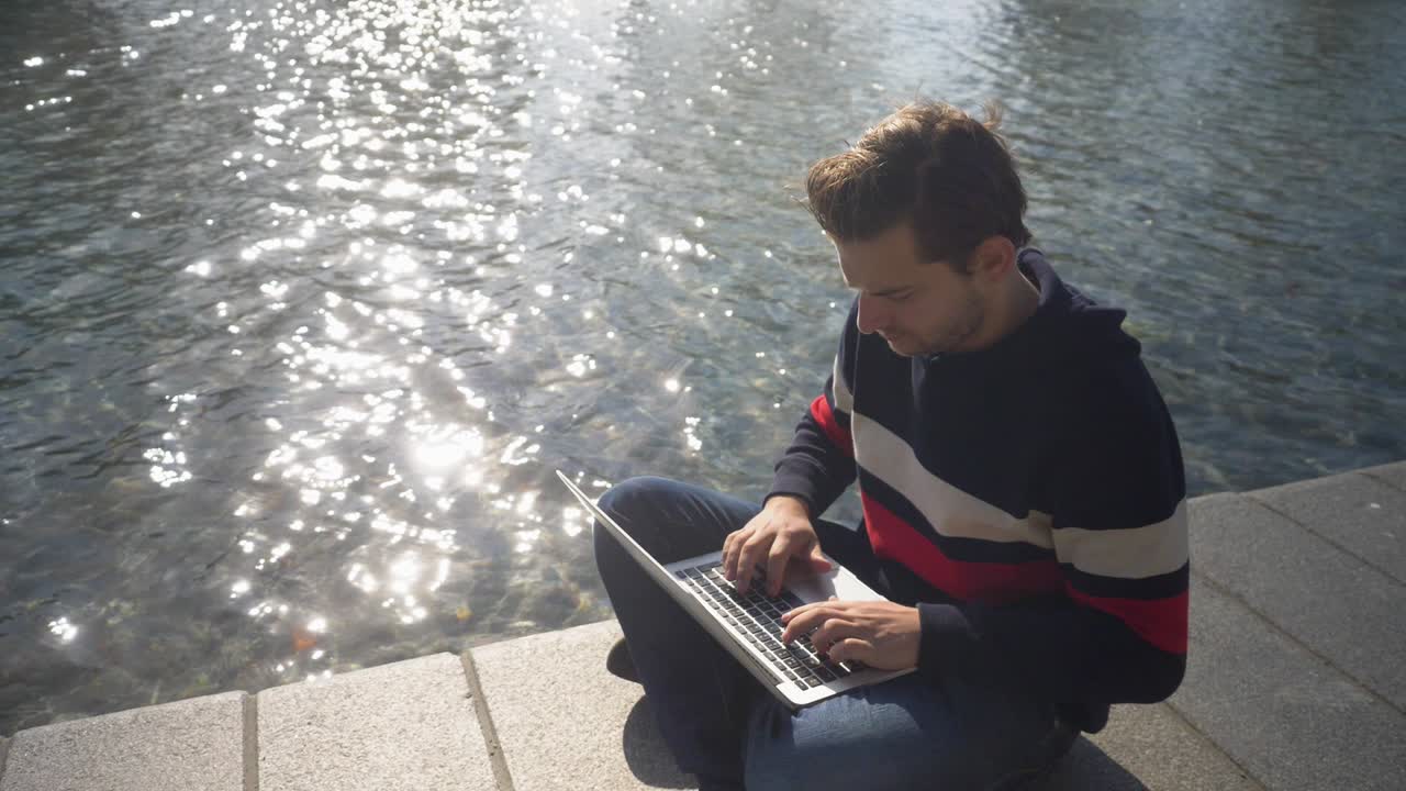 un joven agradable sentado en una orilla del canal en amsterdam escribiendo en el cuaderno