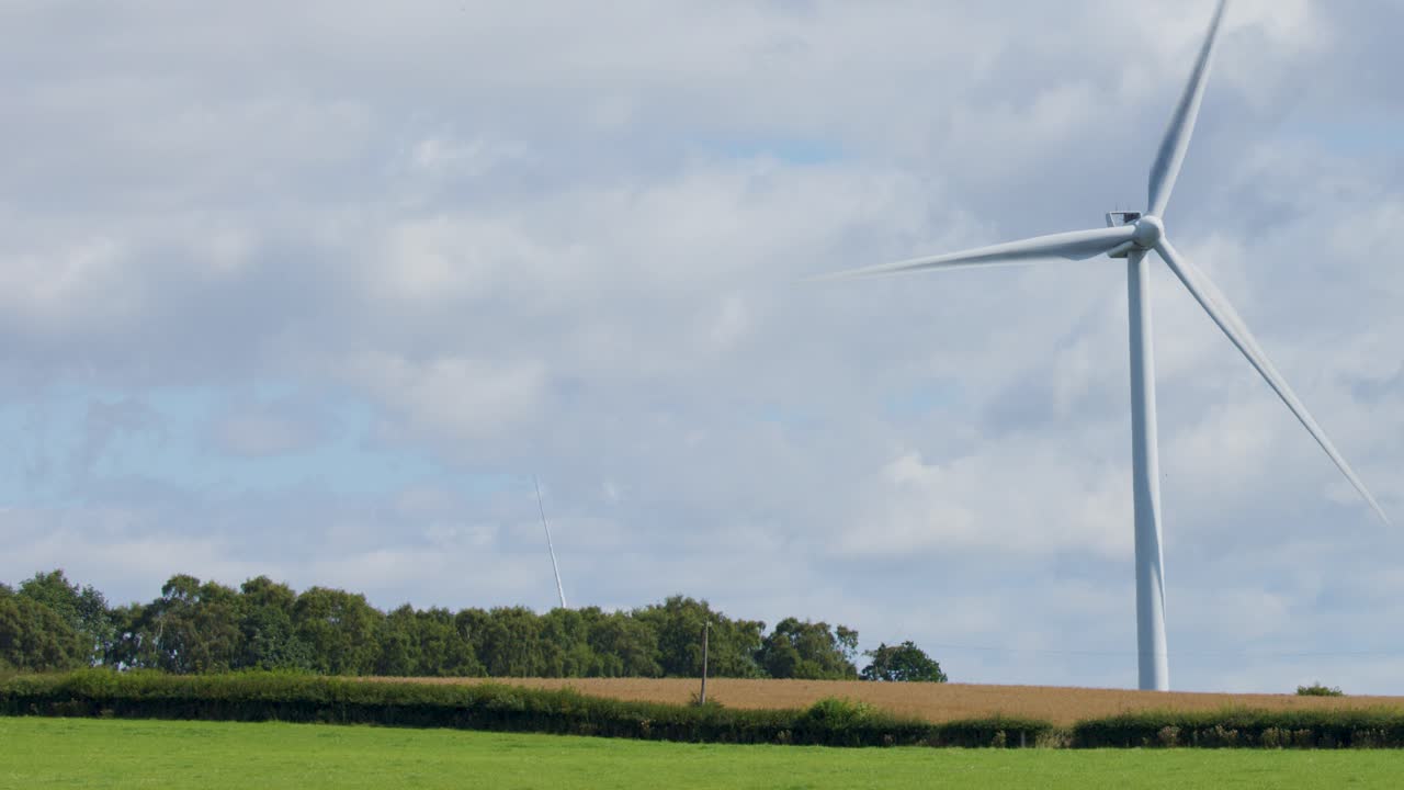 A large wind turbine spins steadily in a rural field near Edinburgh, Scotland. Daylight, wide shot, static camera, partly cloudy sky, green landscape