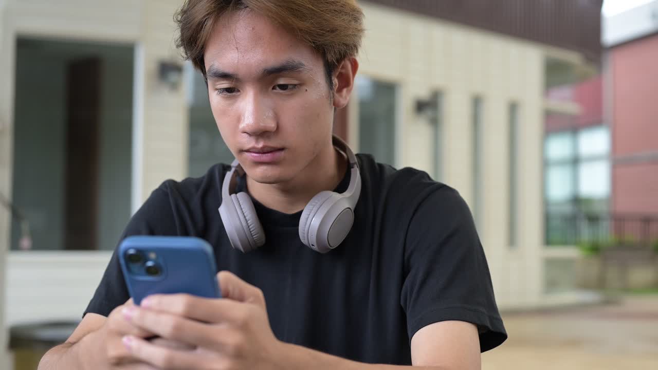Portrait of a young Thai Asian man in casual style sitting and using phone outdoors during summer. Confident, relaxed, and authentic look suitable for various concepts and uses