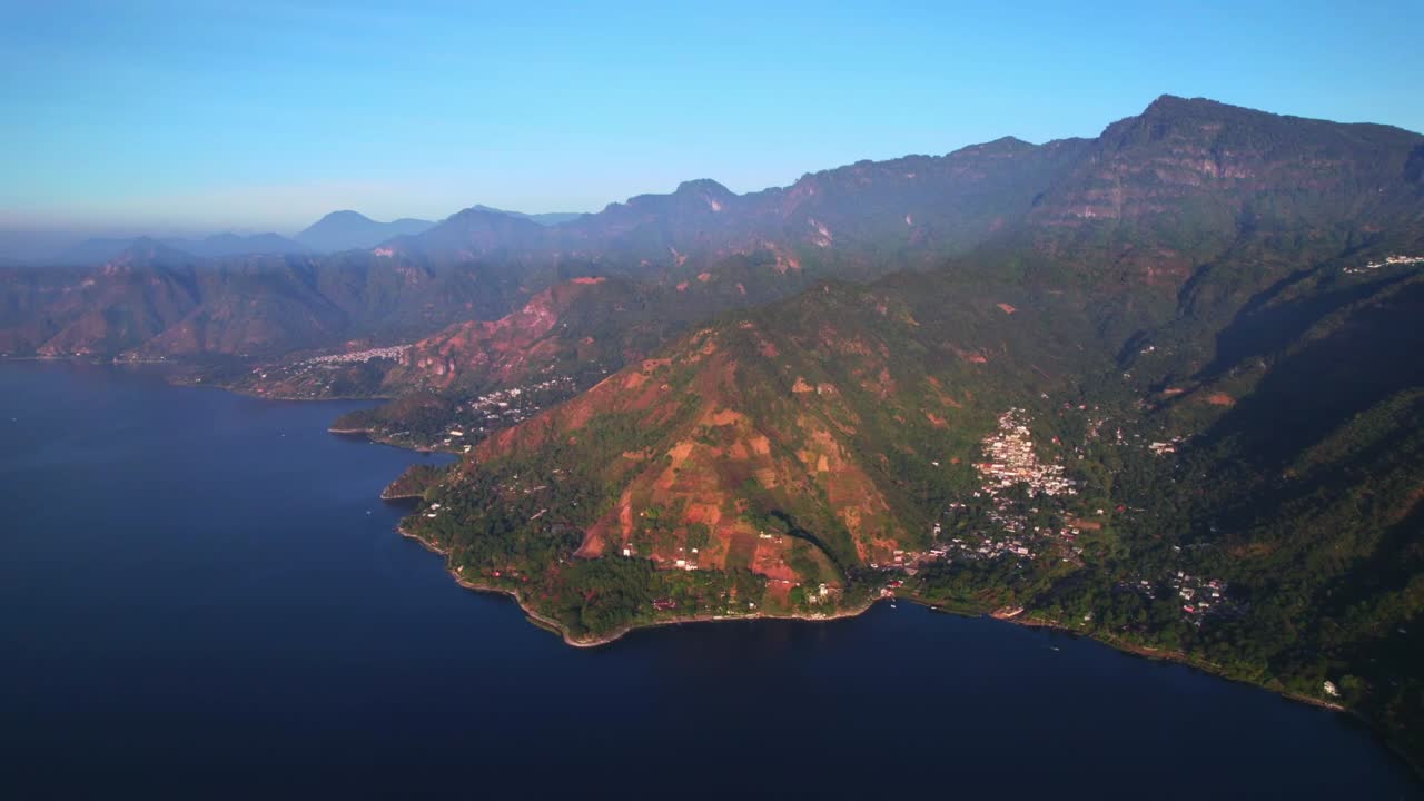 Zoom aereo sulla costa vulcanica del Lago Atitlán, che rivela montagne ripide, villaggi e acqua blu profonda in condizioni di piena luce diurna