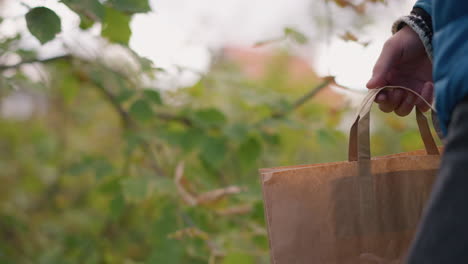 close-up partial view of person in blue jacket holding paper bag while walking, cutting leaves from nearby tree and placing them inside bag, blurred vibrant greenery in background