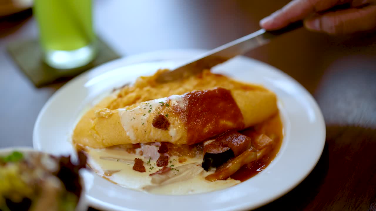 Hand slices omurice with knife on plate, natural light, shallow depth, restaurant dining setting
