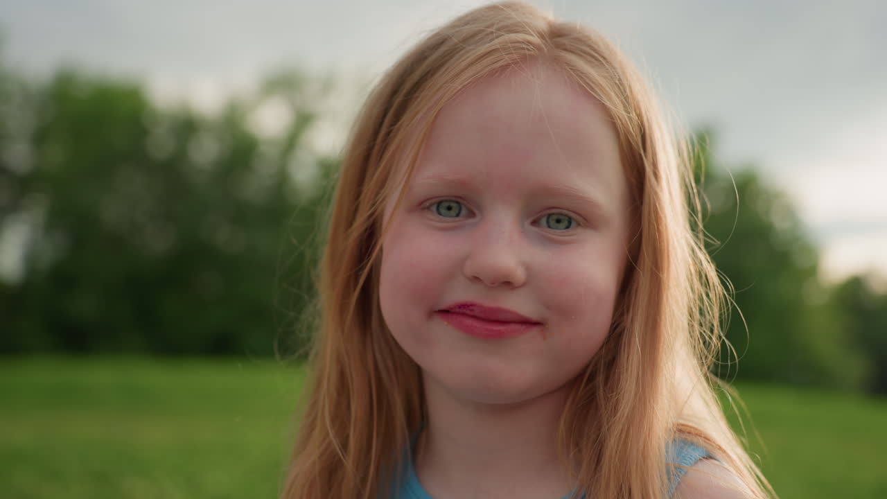 close up white little girl chewing food on green field, flies buzzing nearby, visible sore on lip, serious expression, summer light, natural outdoor scene capturing imperfect childhood snack moment