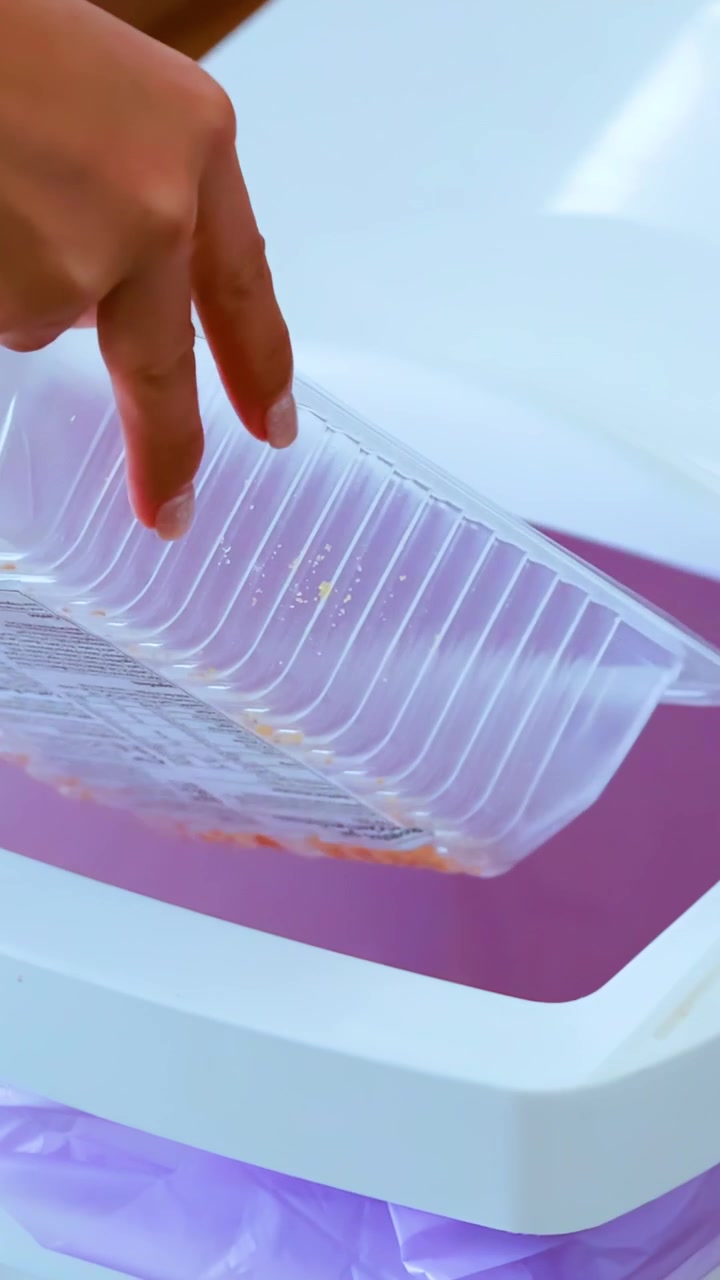 Una persona getta un contenitore di plastica vuoto per alimenti in un bidone della spazzatura bianco rivestito con un sacco viola, in un ambiente interno ben illuminato, chiudendo il coperchio in seguito.