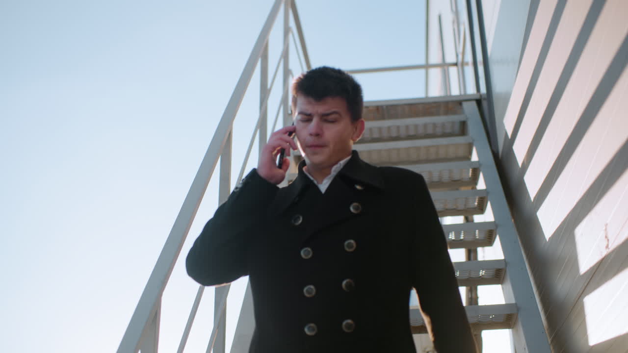 검은 코트를 입은 남자가 전화 통화를 하면서 계단을 내려가는 브리프케이스를 들고, 빛 아래 대화하는 동안 고개를 끄덕이고, 태양빛은 배경에서 밝게 반사됩니다.