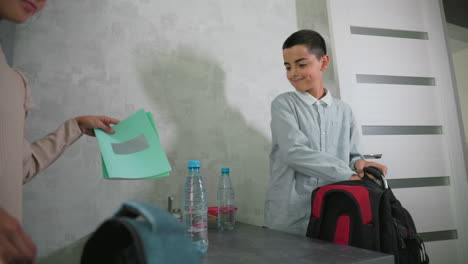 boy in light blue shirt packing black and red backpack for school beside kitchen table with water bottles and books, while girl stands nearby, preparing supplies in morning home environment