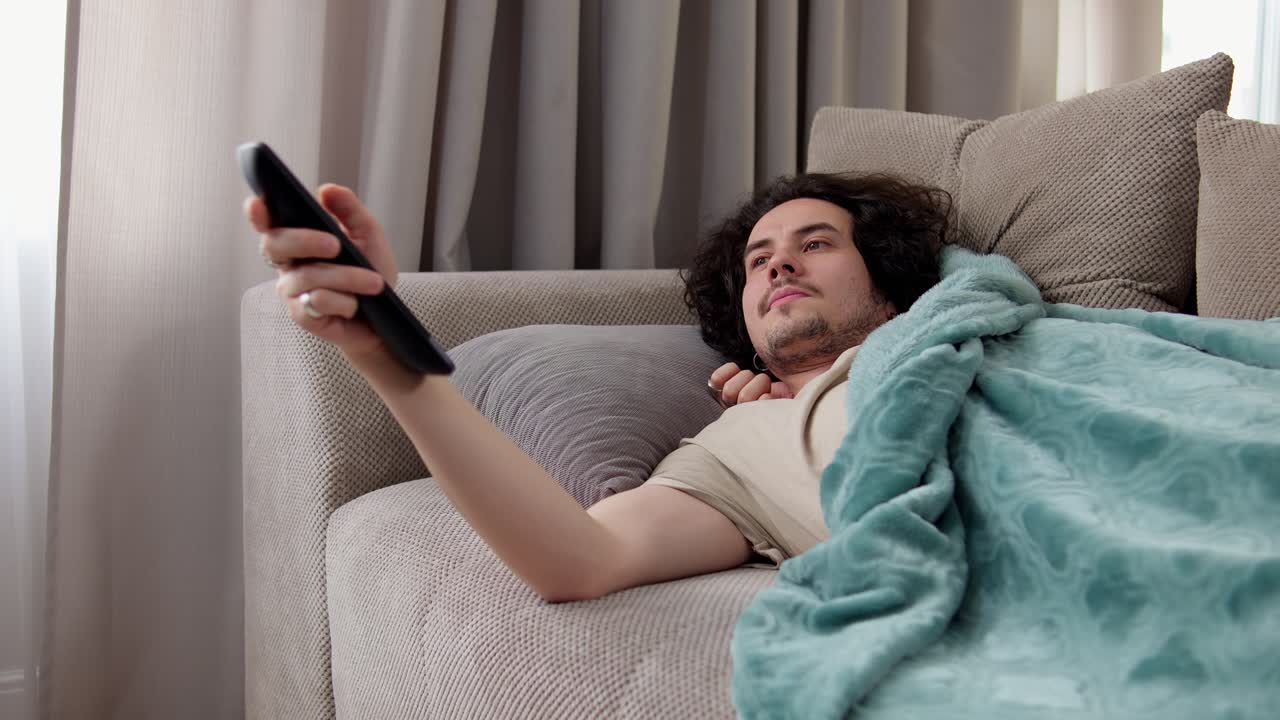 hombre confiado morena con cabello rizado acostado en el sofá se cubrió delante de él y rebobinar canales usando el control remoto en casa