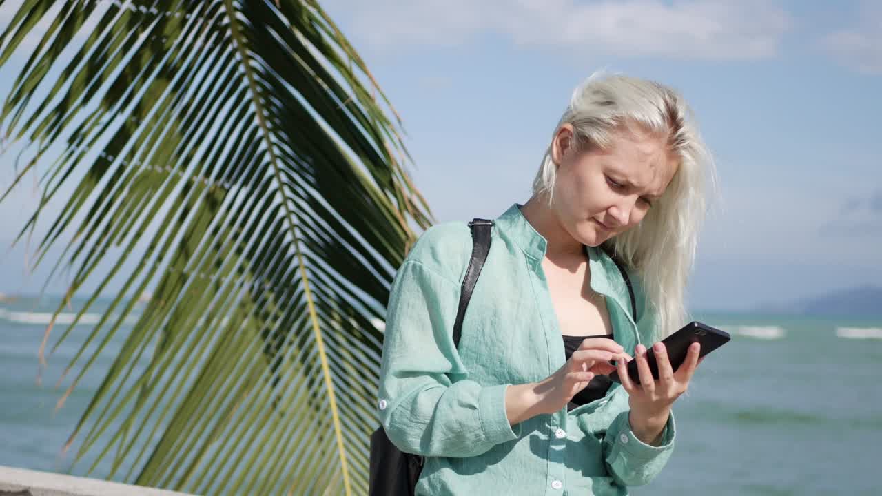 긴 금발 머리카락과 녹색 셔츠를 입은 한 여성이 나무 근처에 서서 배경 바다에 스마트폰을 사용합니다. 소녀 터치 스크린