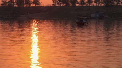 Große-Außenaufnahme-Eines-Sees-Mit-Einem-Lichtstrahl,-Der-Sich-Bei-Sonnenuntergang-Auf-Dem-Wasser-Spiegelt,-Mit-Einem-Treibenden-Boot-Und-Bäumen-Am-Entfernten-Wasserrand