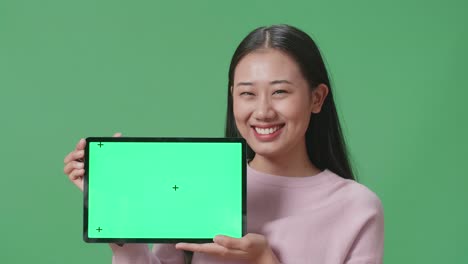 아시아 여성 학생이 녹색 스크린 스튜디오에서 태블릿 녹색 화면 모형을 보여주고 있습니다.