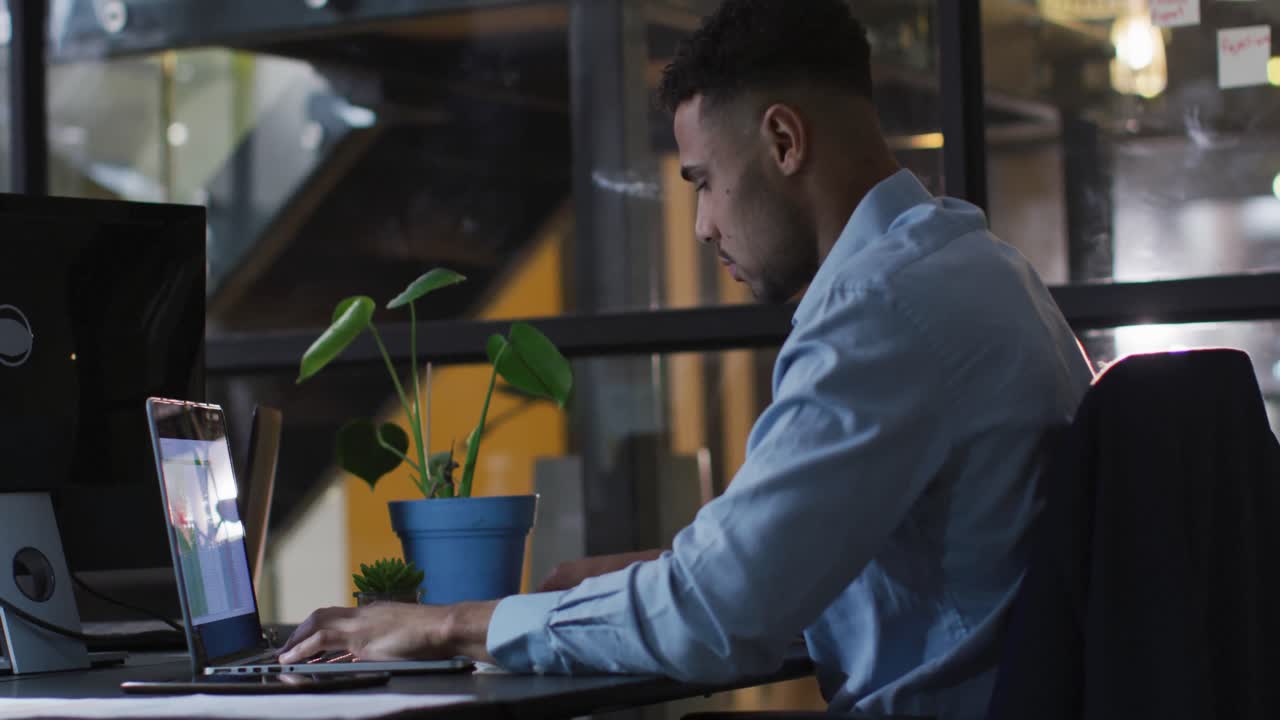 Video of biracial businessman sitting at desk working on laptop at night in office
