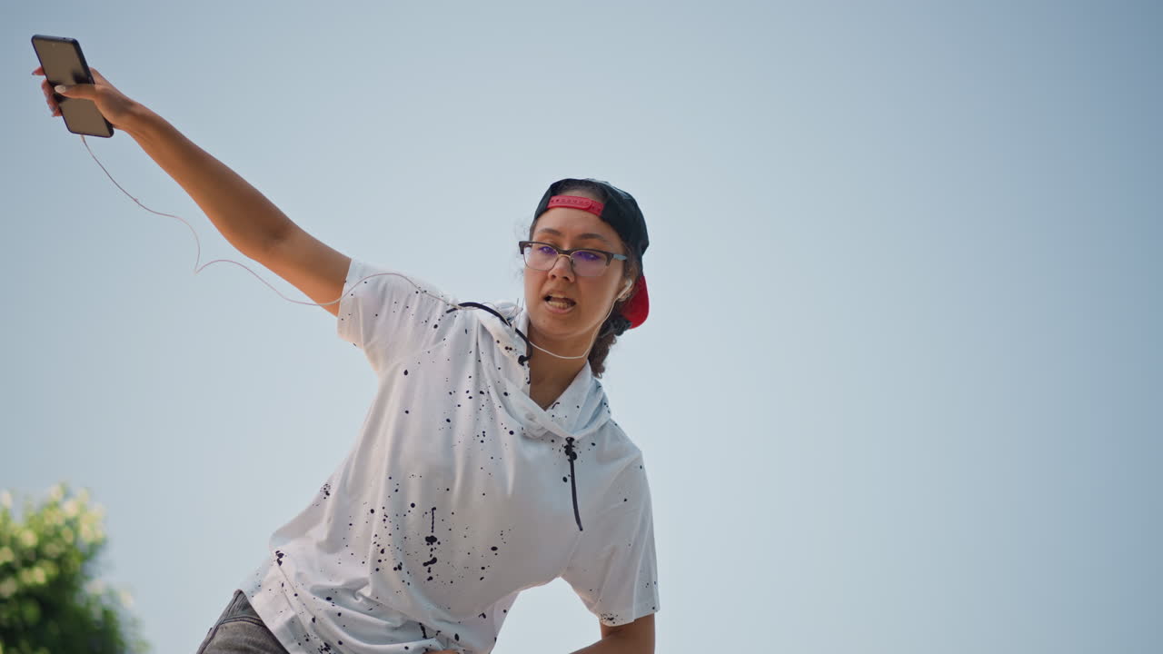 휴대폰을 들고 물을 던지는 십 대, 야외에서 모자와 안경을 착용, 역동적인 로우 앵글 샷으로 맑은 하늘을 배경으로 활기찬 물보라와 움직임을 포착, 이어폰이 늘어진 캐주얼 후드티, 공원