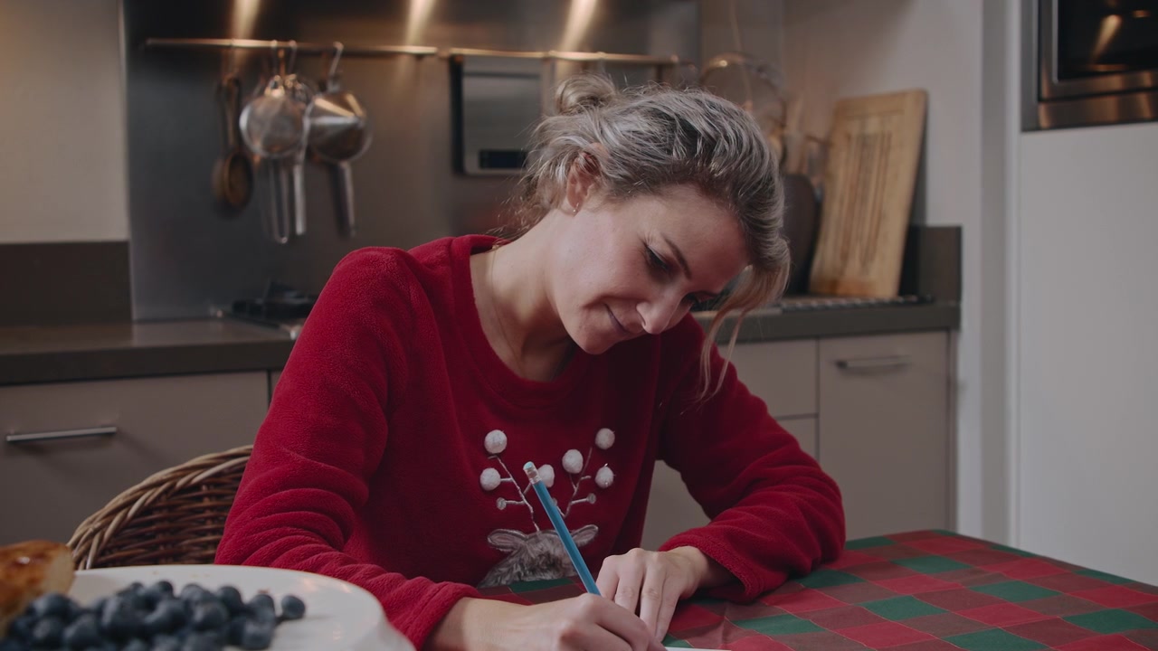 Young caucasian woman writing a Christmas card in her home kitchen finishing and looking at it happily