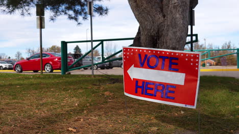 vote here sign pointing left with people driving cars in parking lot background