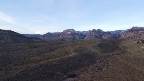 Aerial-shot-of-the-desert-landscape-and-a-dry-riverbed-with-the-Red-Rock-mountains-contrasting-the-landscape-in-the-distance