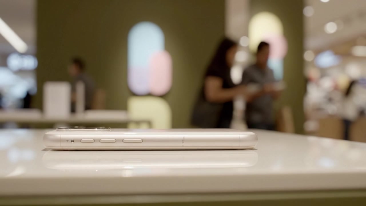 Presentazione di uno smartphone elegante su un moderno bancone di vendita al dettaglio, evidenziato da uno sfondo contemporaneo con superfici lucide e acquirenti sfocati in un ambiente di negozio