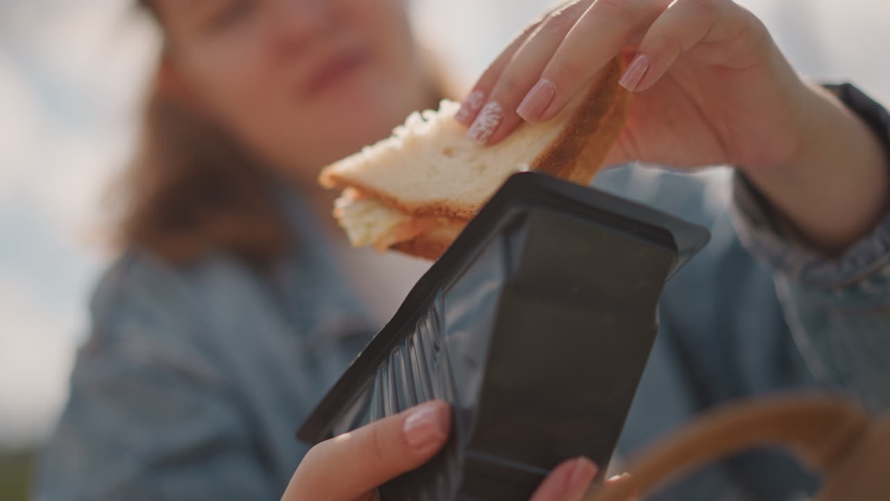 Young white woman opening sandwich box briskly, denim jacket, sunlit field, closeup hands pulling black container lid, revealing toasted sandwich with jam, candid snack moment, relaxed picnic vibe