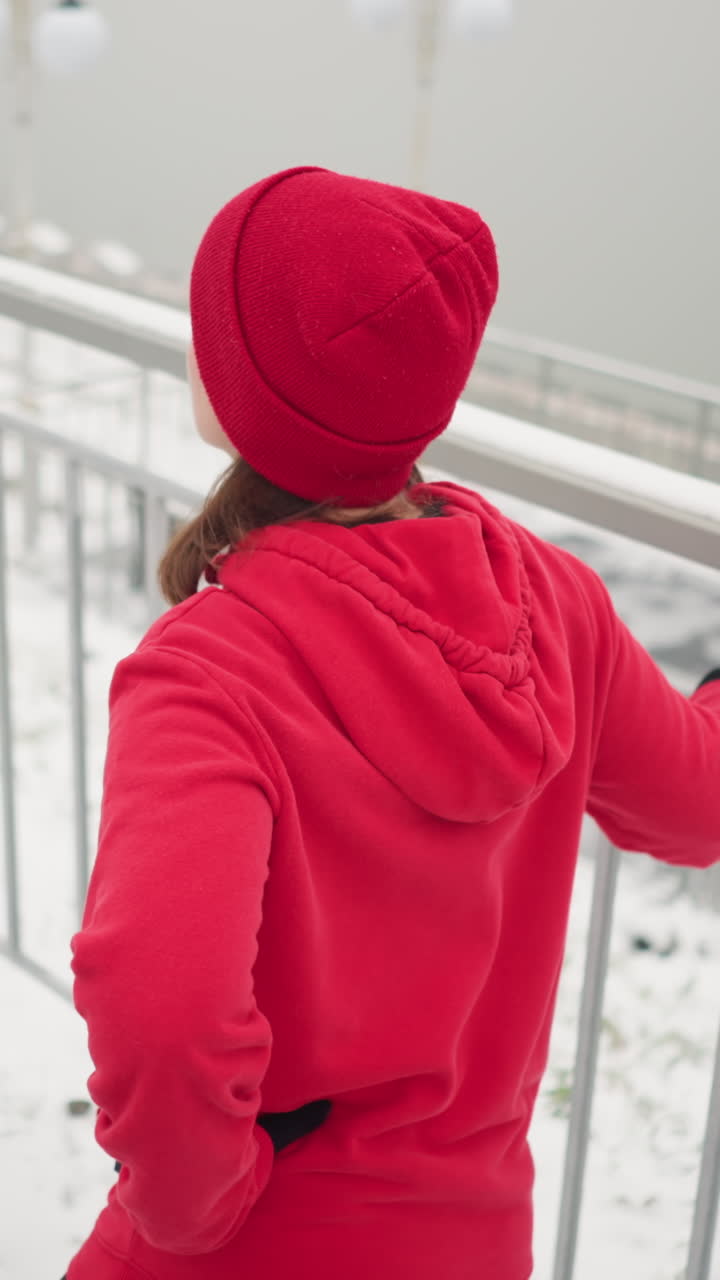 business woman exercising outdoors during winter holding iron railing for balance stretching leg back and forth with warm smile surrounded by snowy ground foggy atmosphere river bridge light poles