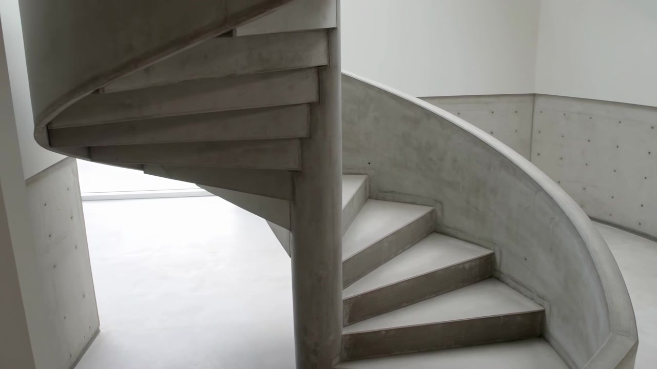 Curved cast concrete spiral staircase viewed from mid-height in a bright minimalist interior. The winding steps and smooth balustrade wrap around a central