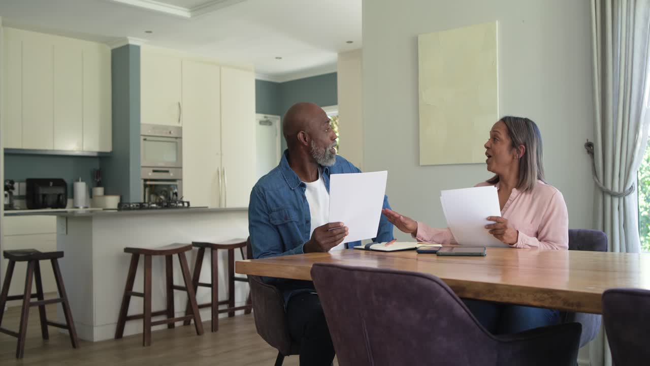 With pages, diverse couple sitting at dining table, reading, tapping tablet to cross check bills