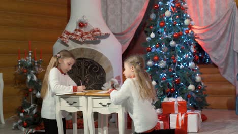 Two sister girls in white sweaters draw in the house with New Year's decorations, gifts and a Christmas tree. Log country house with a fireplace