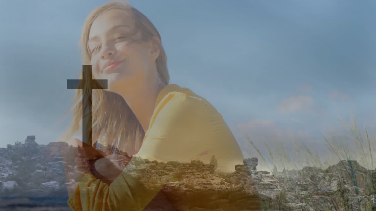 Smiling woman holding cross symbol against rocky landscape under blue sky