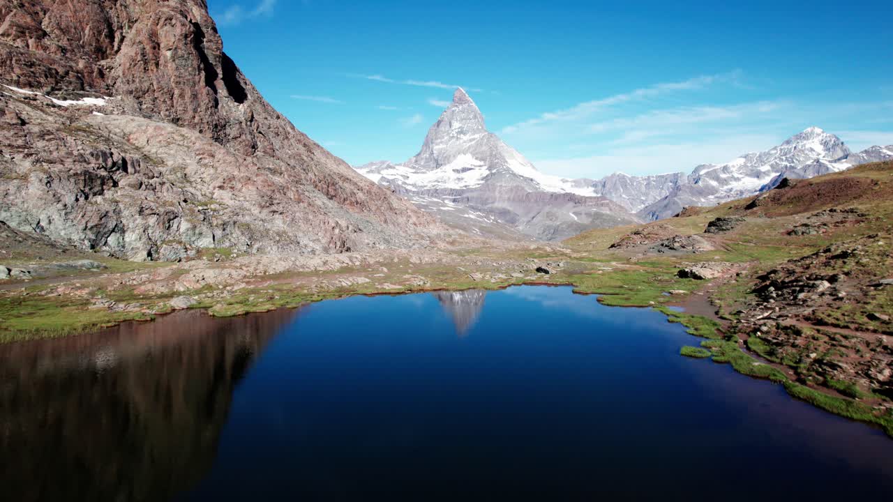 Filmische Luftaufnahme, die die Spiegelung eines Gletschersees des ikonischen Matterhorns enthüllt, das über dem Zermatter Tal thront. Unberührte Alpenlandschaft mit felsigem Gelände und klarem blauem Himmel in den Schweizer Alpen.