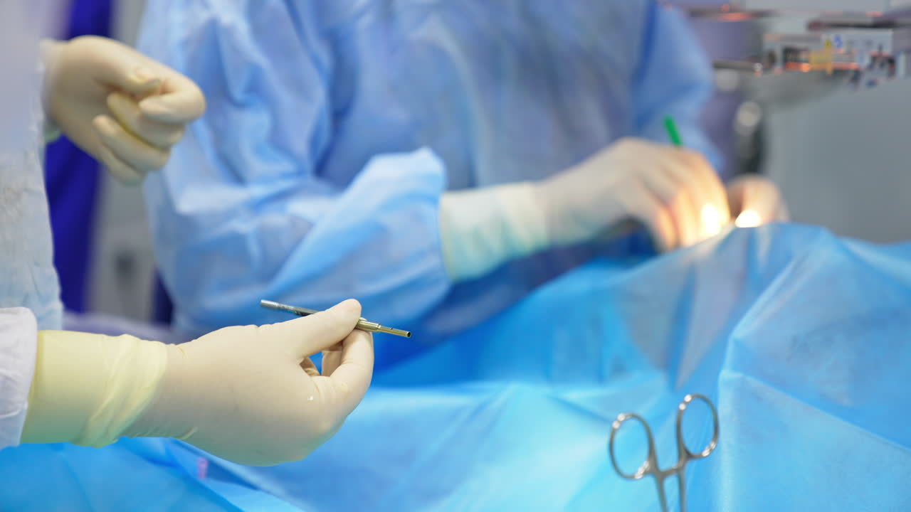 Nurse's gloved hand passes thin instrument to a chief surgeon. Close up. Doctor working under equipment in blur at backdrop.